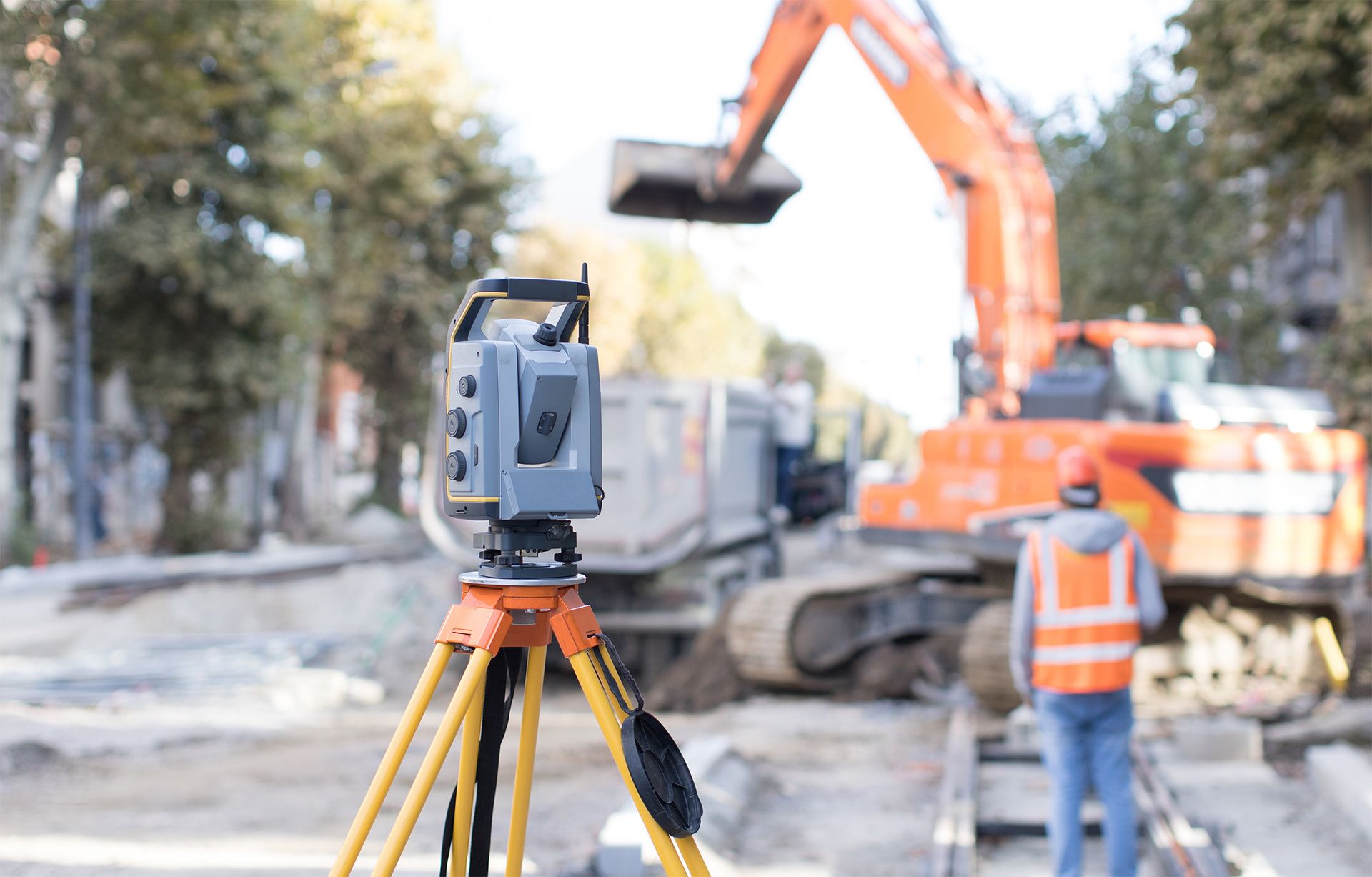 Tachymeter im Einsatz auf einer innerstädtischen Baustelle, im Hintergrund ein Vermessungstechniker und Baggerarbeiten